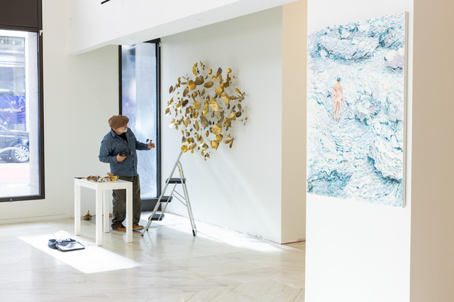 A person wearing a hat and denim jacket arranges a gold and brown leaf sculpture on a white wall, with a table, ladder, and blue ocean-themed painting nearby.
